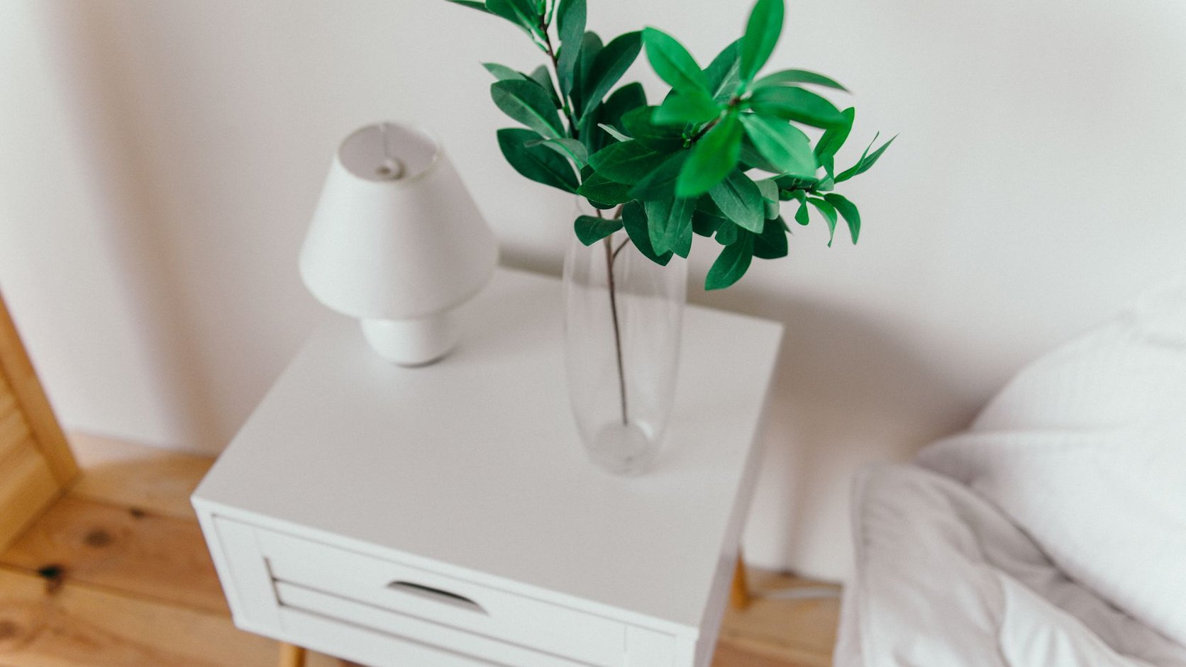 Mesa lateral branca com luminaria pequena e vaso de folhagem em ambiente suave